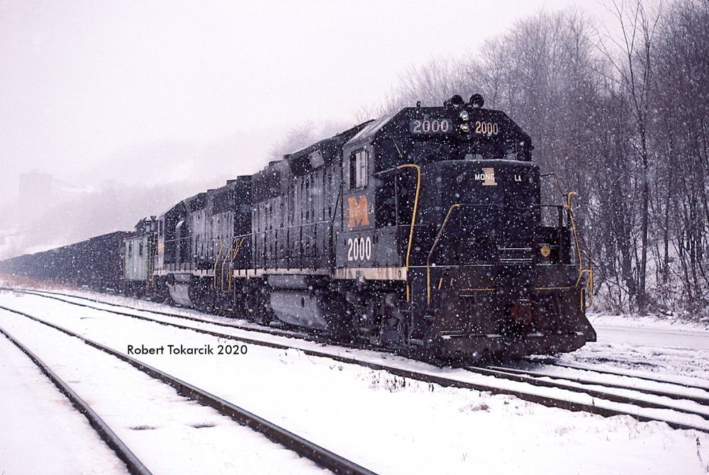 two black diesel locomotives push a green caboose and a line of hopper cars from behind, in the midst of a heavy snow