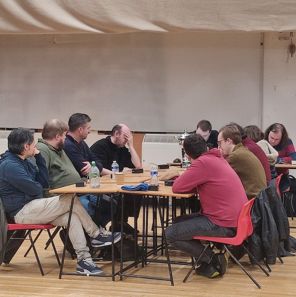 Four men sit on the left of a table, three men and a woman sit on the right. It's a school assembly hall. Iain Thoms holds his head in his hands as they think about quiz questions.