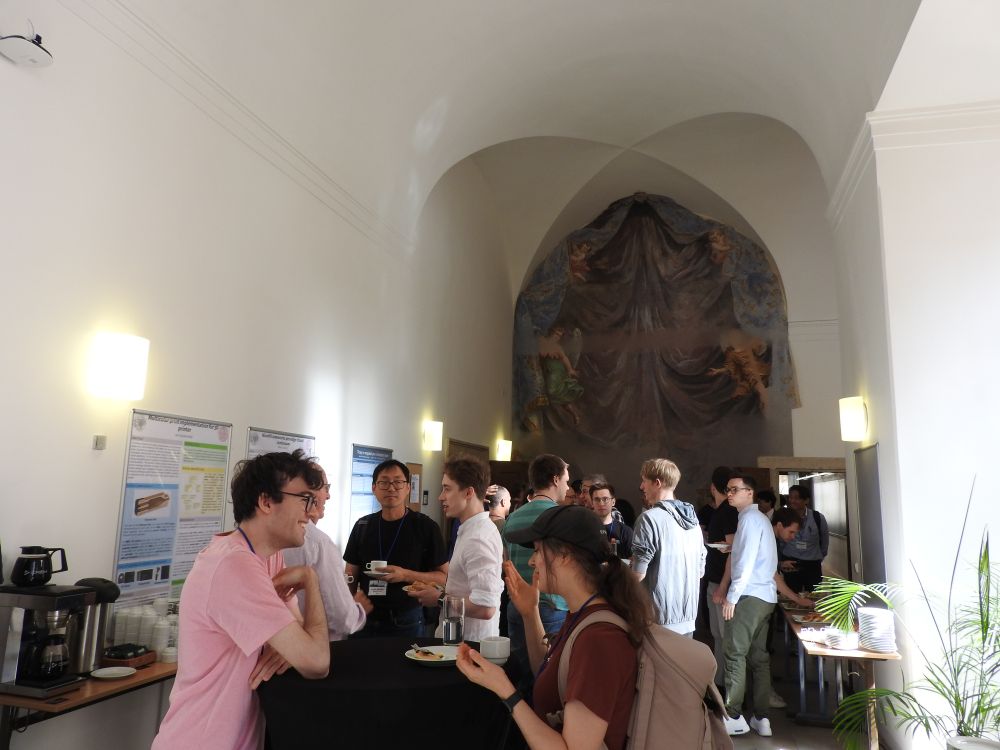 A photo of a coffee break at <Programming> 2025. The room has a high ceiling and the wall in the back has a big painting on it. In the foreground, people are discussing and eating snacks.