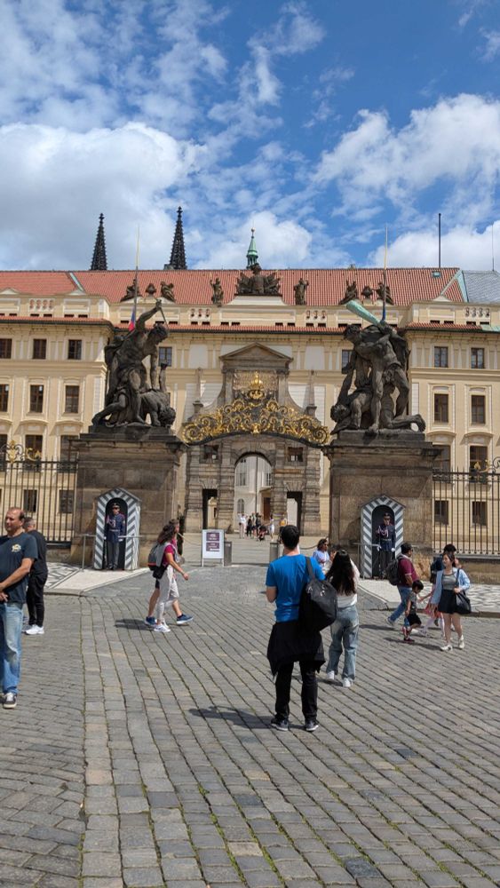 A photo of the gate near Prague Castle. Some people are standing in the foreground. In the middle of the photo is a gate with intricate decorations and two guards posted at the sides. There is an orange-ish building behind the gate. Behind the building, the tops of two antique castle towers can be seen.