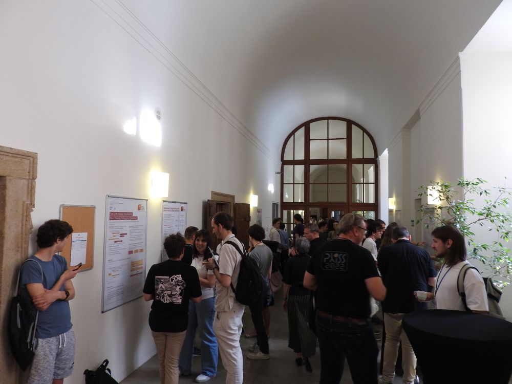 A photo of the first round of the student research competition at <Programming> 2025. There is a hallway with 7 SRC posters pinned to the walls. Groups of people are standing around the posters; the students explaining their research to the audience.