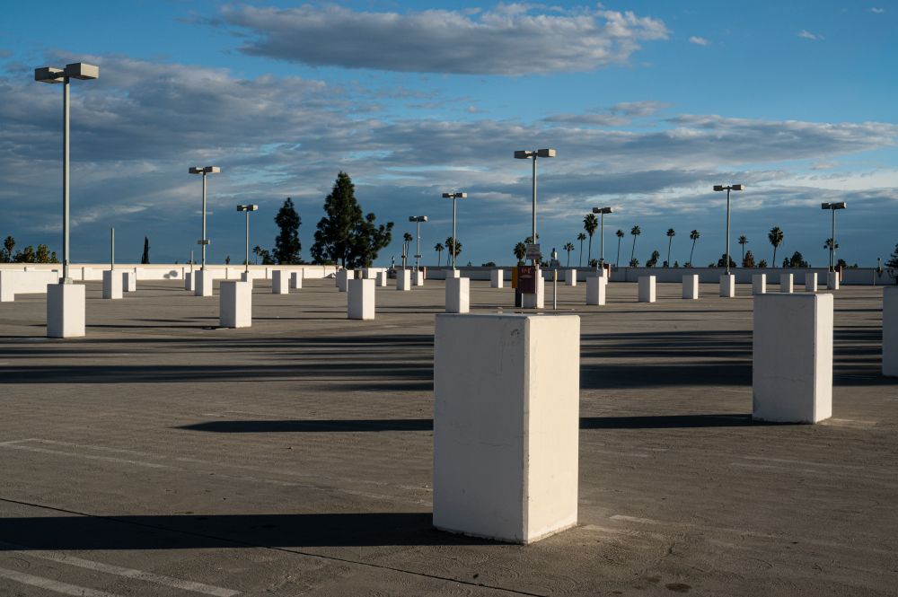 Image of a parking lot with bollards, light poles and trees all creating layers upon layers of repeating lines. As clouds float in the background creating their own lines and layers. Not a car nor human to be found in the image