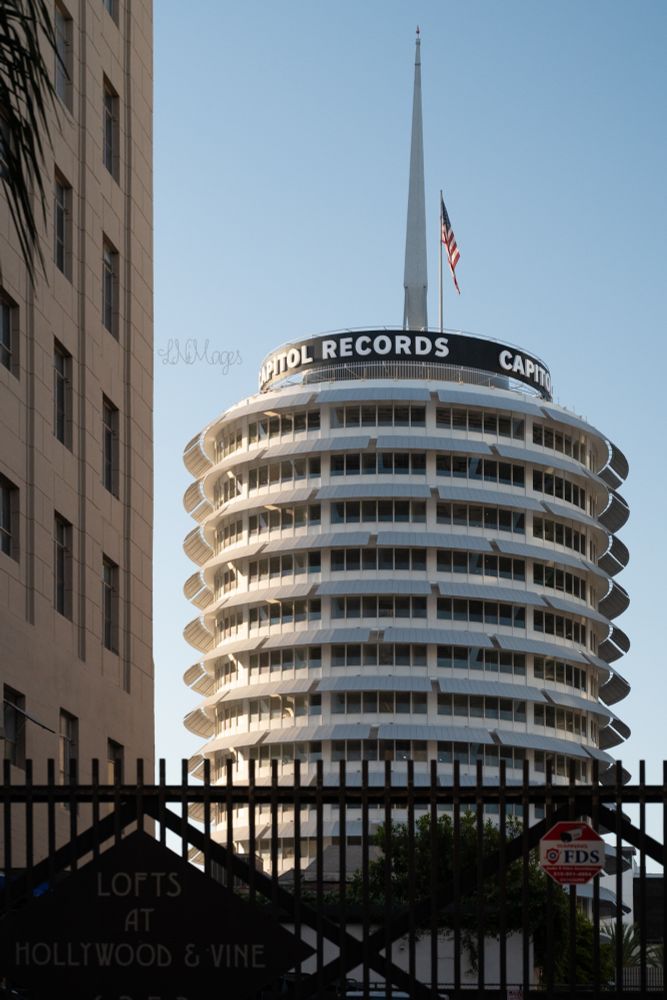 Image of the capitol records building behind a fence that says lofts at Hollywood & vine. An iconic place in Hollywood CA. The building has been used in tons of movies and TV shows. Including when the aliens arrived in Independence Day and people were gathered on top to greet them. The white round building is recognizable anywhere 
