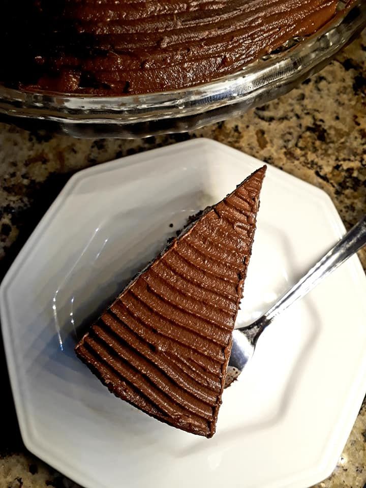 Fotografia colorida. Fatia de bolo de chocolate coberto de ganache, em um prato branco octogonal de porcelana, com um garfo de sobremesa. Na parte superior da imagem, parte do bolo é vista, em um prato de vidro. O fundo é uma bancada de granito amêndoa.