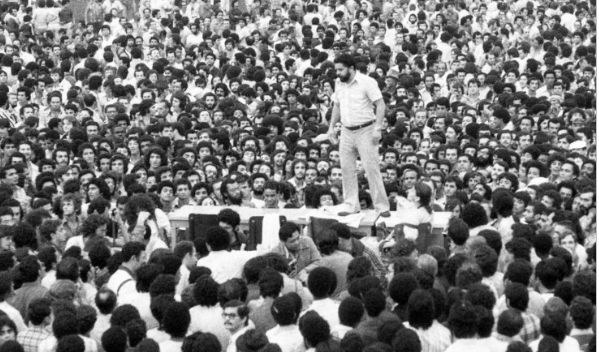 Fotografia PB. Lula em pé, em um plataforma elevada, rodeado por trabalhadores, na greve geral de 1979. Crédito: Fernando Pereira. Acervo: Centro Sérgio Buarque de Holanda/Fundação Perseu Abramo.