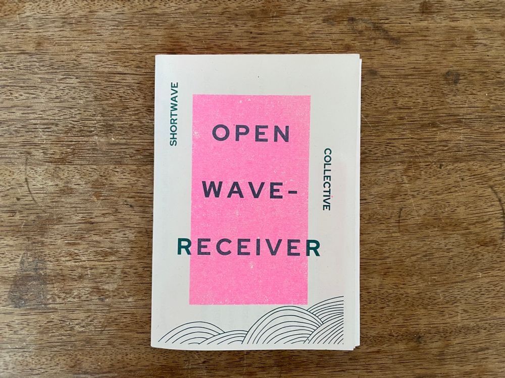 A zine on a wooden table - waves rise up the bottom of the image, a hot pink rectangle in the middle, stamped across the top the words Open Wave Receiver / Shortwave Collective
