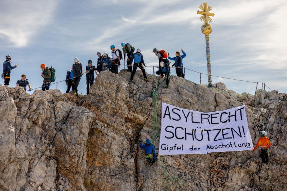 Eine Gruppe Leute steht auf dem Gipfel der Zugspitze. Zwei von ihnen haben sich mit einem großen Transpi abgeseilt, auf dem steht: "Asylrecht schützen! Gipfel der Abschottung".