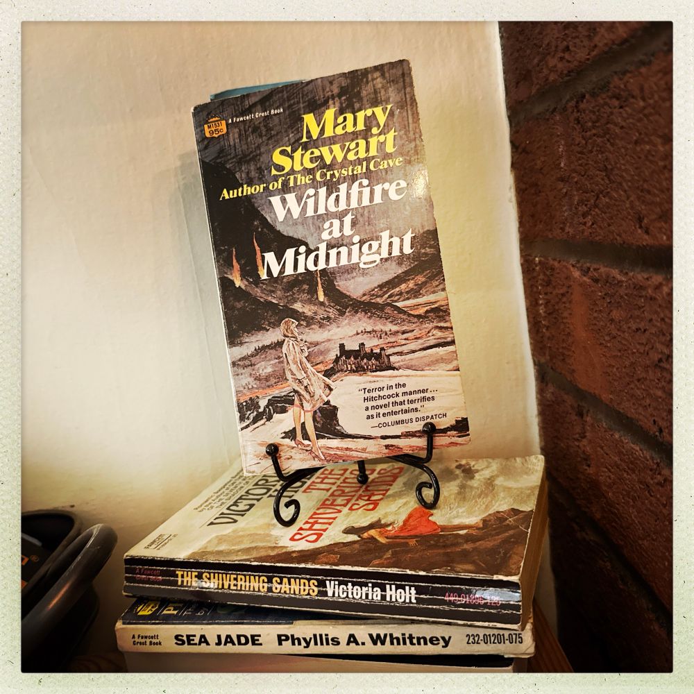 Stack of c. 1960s suspense paperbacks including Mary Stewart’s Wildfire at Midnight with a woman in a coat at the top of a hill with burning fires and a large house in the background. 