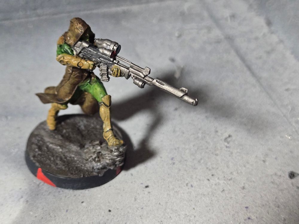 A Hassassin Lasiq stepping forward on a small rock, aiming her sniper rifle forward. She has light brown armor, green fatigues, and a leather brown coat.