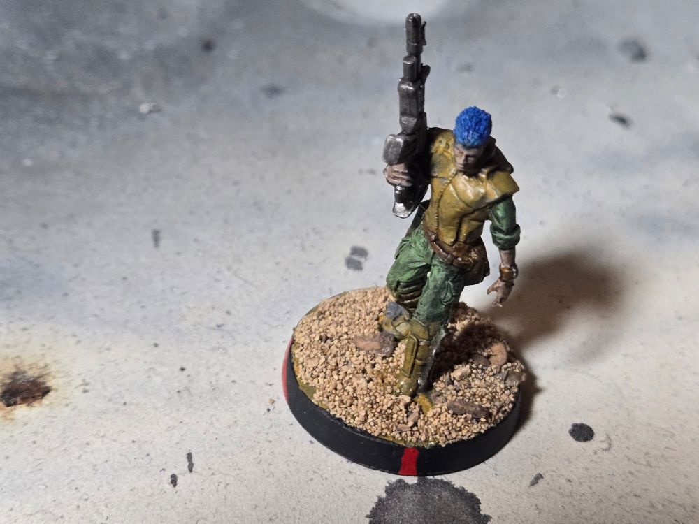 The front of a Daylami. Male with blue hair and holding a rifle in his right hand, barrel pointed up. His armor is light brown over green fatigues.