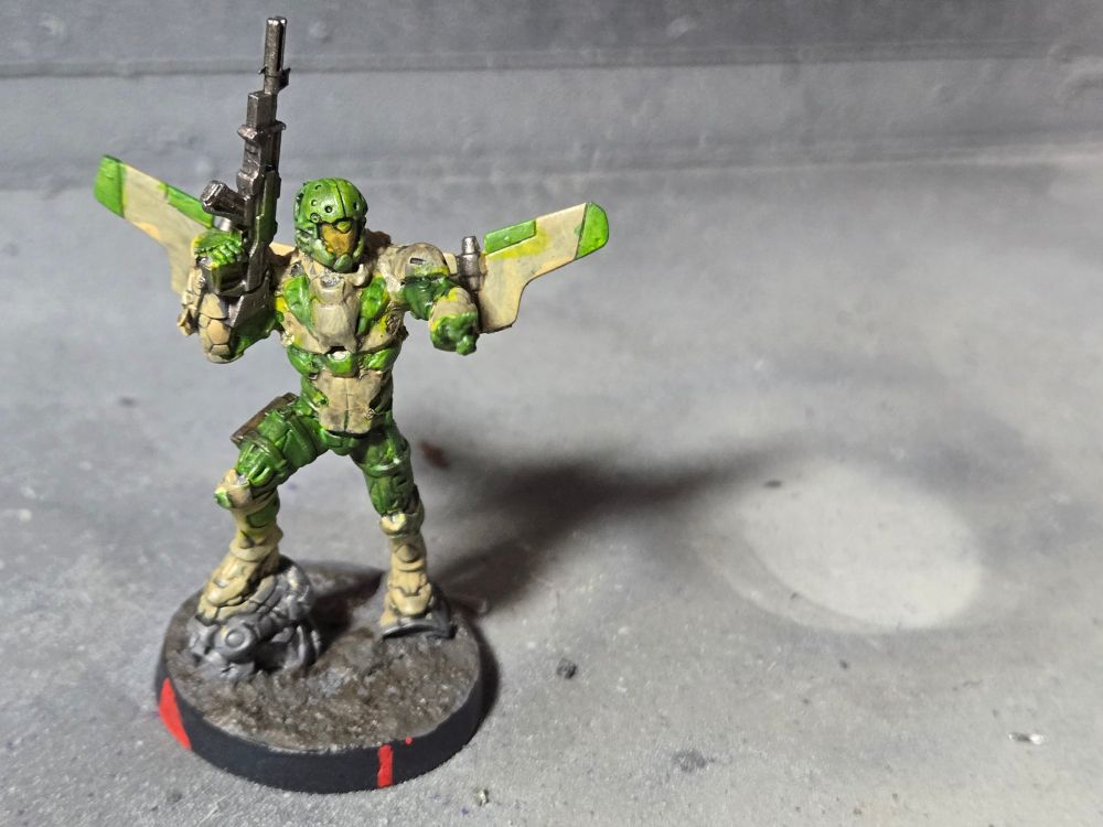 A Hassassin Ragik hacker. Standing upright with his right foot on a tactical rock. His right hand holds his rifle pointed straight up. His left had is held out i n the hacker hand pose. His armor is sand colored with green accents over green fatigues. His helmet is green with a yellow face plate. The wings of a jet pack, also sand and green, are sticking out to the sides.