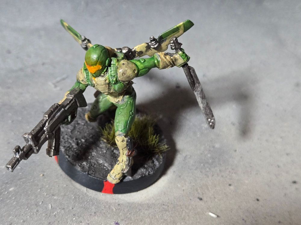 Hassassin Ragik landing and aiming forward with a rifle in his right hand and holding a sword out behindhim in his left hand,  bladepoint down. Armor is light sand and light green. Helmet is light green with an orange face plate.