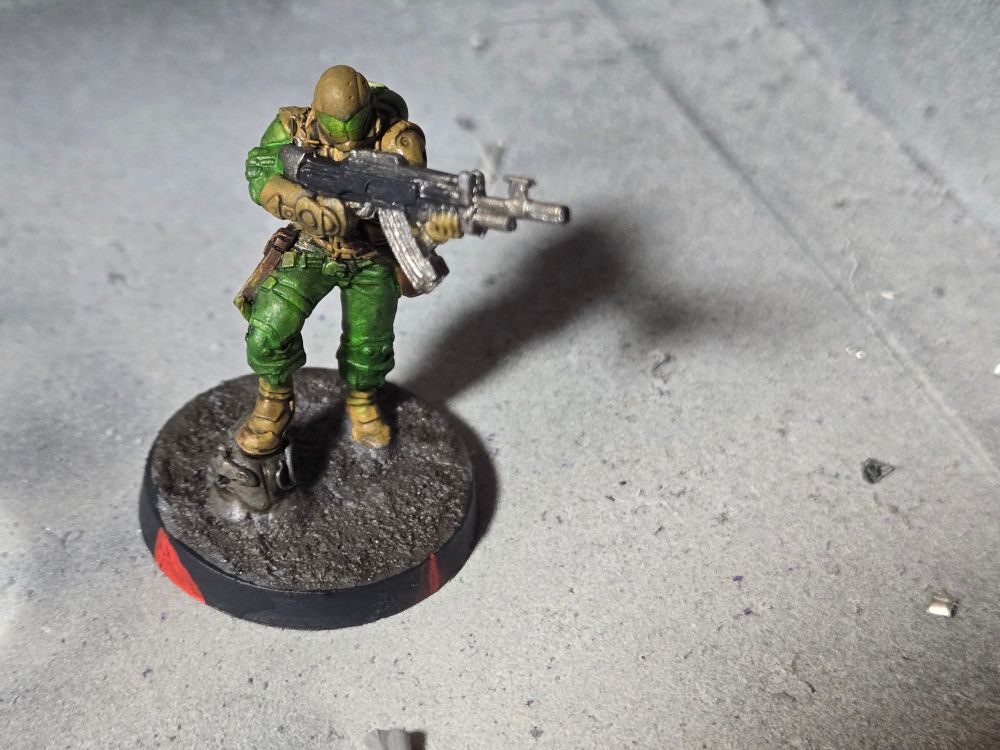 A Hassassin Muyib with his right foot up on a small rock, aiming his rifle to his left with both hands. He has light brown armor over green fatigues, light brown boots, and a light brown helmet with a green visor covering his face. A leather brown holster is on his right leg.
