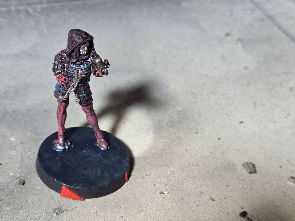 Fixer wearing a thigh length dark purple jacket with blue accents and a hood over her head. She has pink leggings, gloves, and shoes. She wears a silver mask and holds cylindrical metallic device in her left hand.