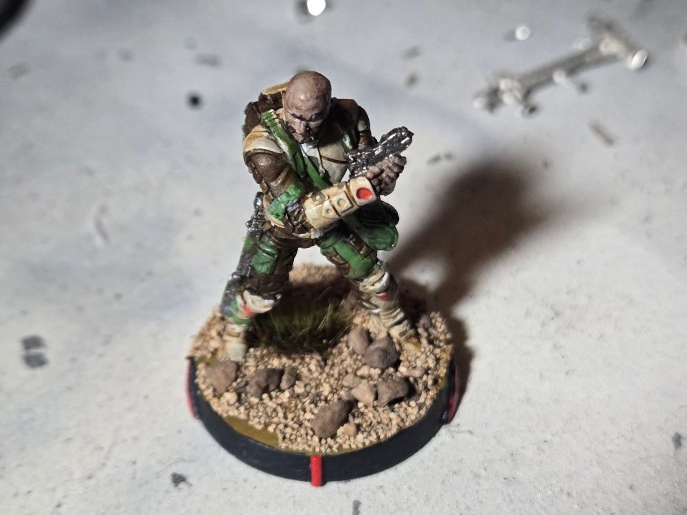 Front of male Ghulam doctor holding a medkit.  Light brown skin, bald, black facial hair. Dark brown uniform with green belts and pouches. Light tan boots and armor.