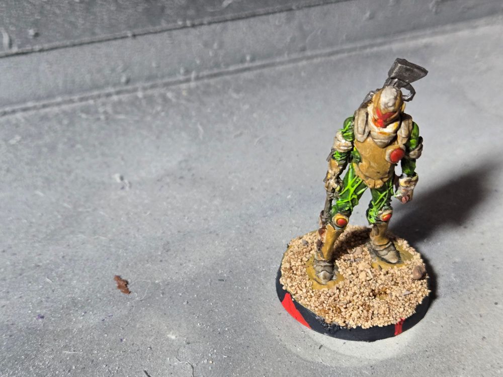 A Hassassin Fiday standing upright with arms at his sides, head and shoulders turned slightly to the right. There is a knife sheathed on his right boot and the but of a rifle can be seen over his left shoulder. His armor is brown and tan over a green undersuite. His face plate is red.