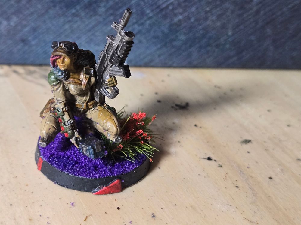 A female Hunzakut in light tan fatigues squatting over purple sand next to some red flowers. She's holding a rifle up in her left arm and placing a deplorable object on the ground with her right hand.