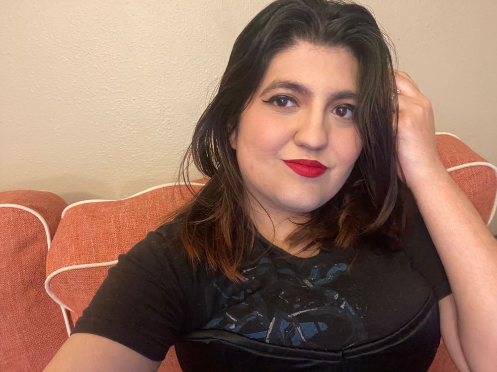 a selfie of anya on a pink couch. she is wearing victory red lipstick & a black panther t shirt under a silk corset. they are smiling at the camera.