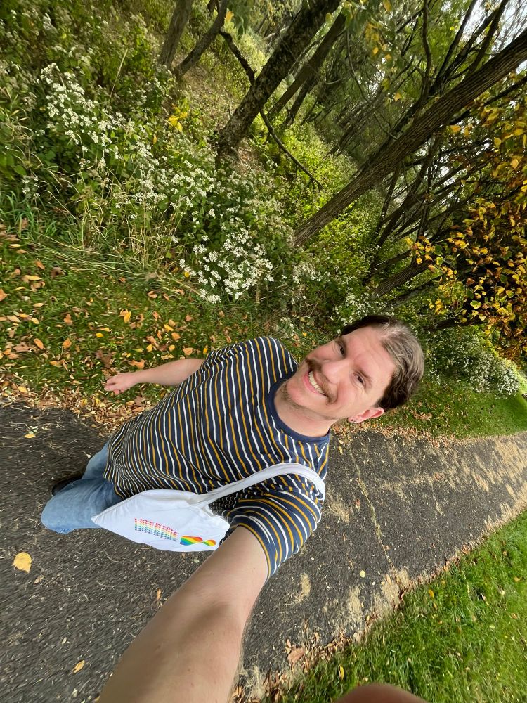 Grinned whiled walked by the trail sidewalk at the national park. Queer behr wore a striped shirt and jeans. Shoulder bag. 