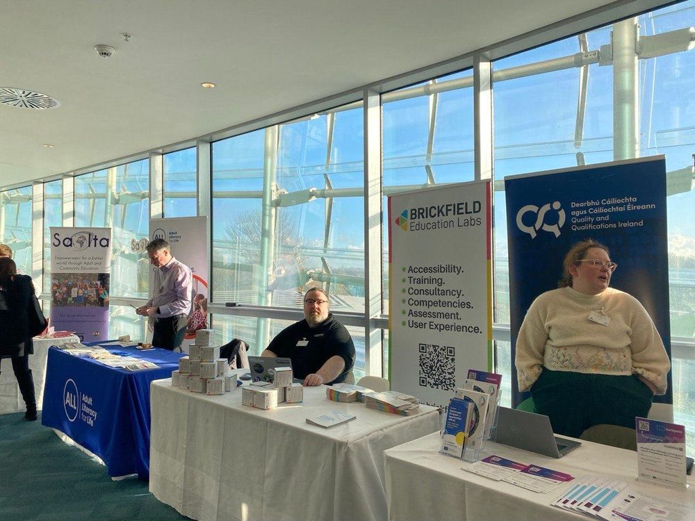 A bright, glass-walled conference space with several exhibitor tables lined up. People stand or sit behind the tables, displaying brochures, laptops, and stacks of small boxes. The Brickfield Education Labs stand features a large banner highlighting accessibility, training, consultancy, and user experience. Taken at the AONTAS Adult Education Summit.