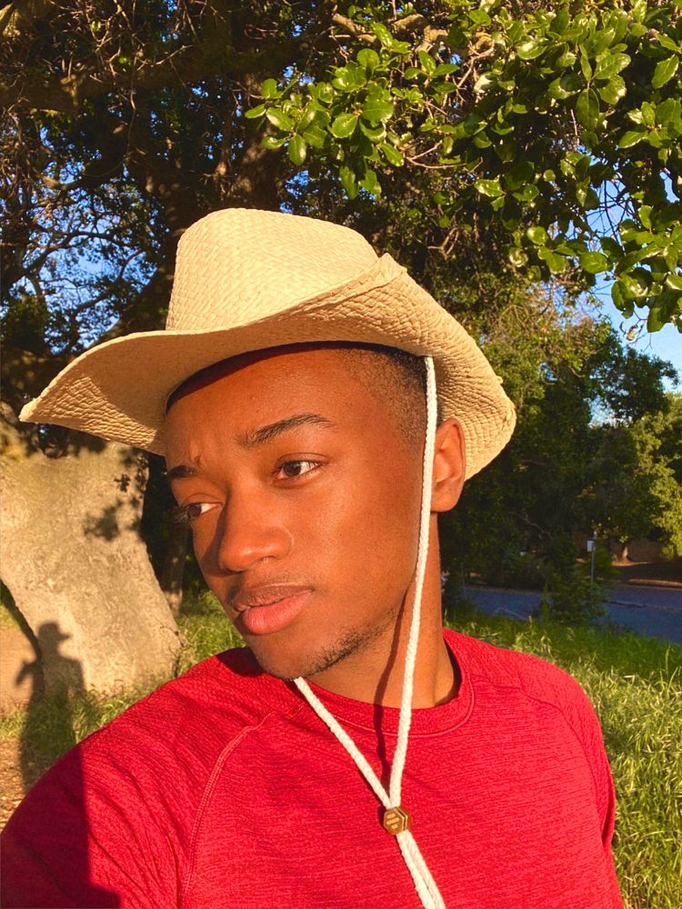 me taking a selfie. i’m standing in front of a big tree and some grass and wearing a red shirt and a tan straw cowboy hat.  
