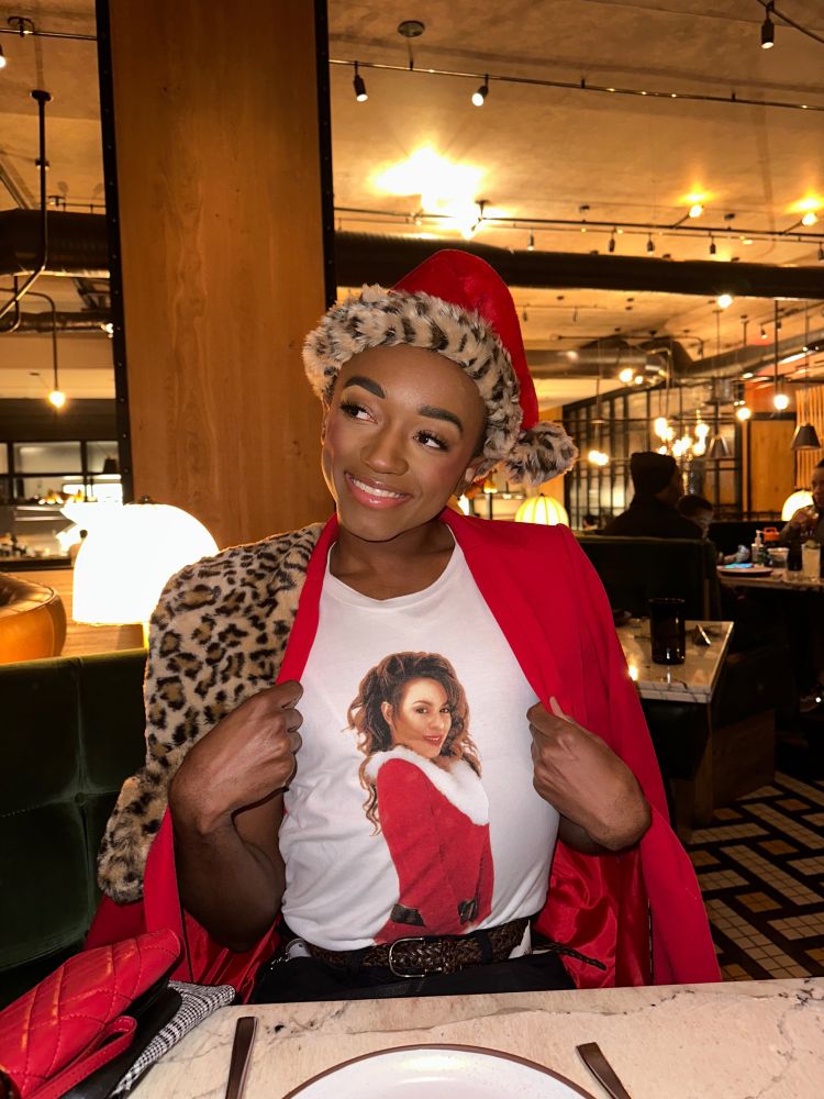 me posing for a photo in a restaurant. i’m smiling and tilting my head as i look off to the side. i’m wearing a white t-shirt with an image of mariah carey in a santa outfit and a red jacket over my shoulders, as well as a cheetah-print adornment over my shoulder and a santa hat with a cheetah-print brim. 