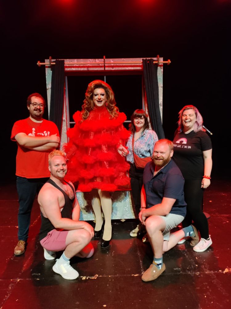 Group meet and greet photo on stage with drag artist Kate Butch. Josh, James, Zoe, Stuart and Rosie are variously stood or kneeling around Kate, centre state, elegant red dress, big brunette wig, gorgeous. Everyone smiling. 