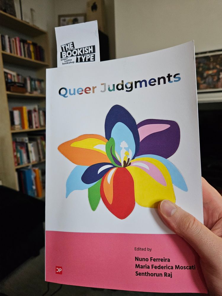 Picture of James' hand holding the Queer Judgments book, edited by Nuno Ferreira, Maria Federica Moscati and Senthorun Raj. It features a graphic design image of a queer flower, with petals each being a colour from the progress pride flag