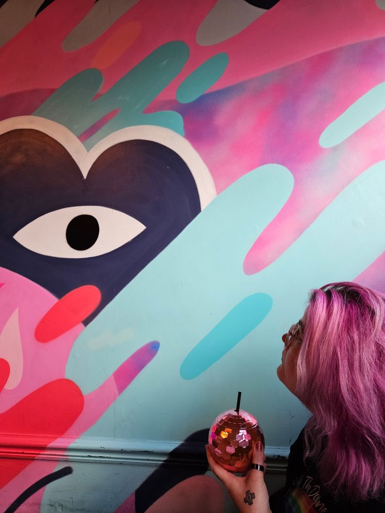 Beautiful tableau of Rosie, with her pink hair and pink disco ball cocktail in hand, looking up in profile at the wall behind, which features pink and blue dubs of graffiti 