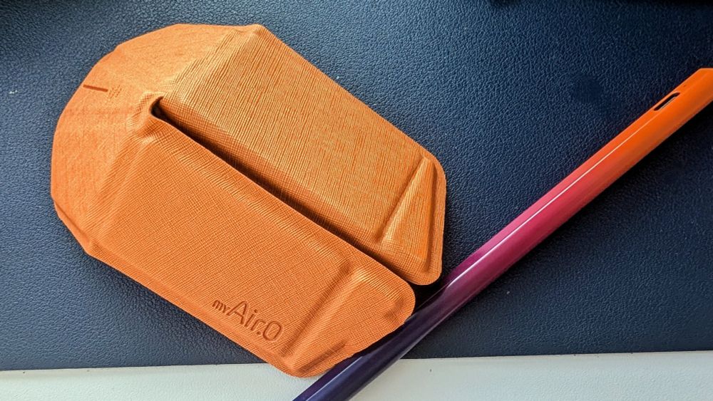 Image of white desk with navy blue desk mat. Pointed towards the top left corner is a metallic orange computer mouse with "myAir.0" logo emblazoned on the bottom. Pointed towards the bottom left corner is a multicolored (orange, pink, purple) Apple stylus. 