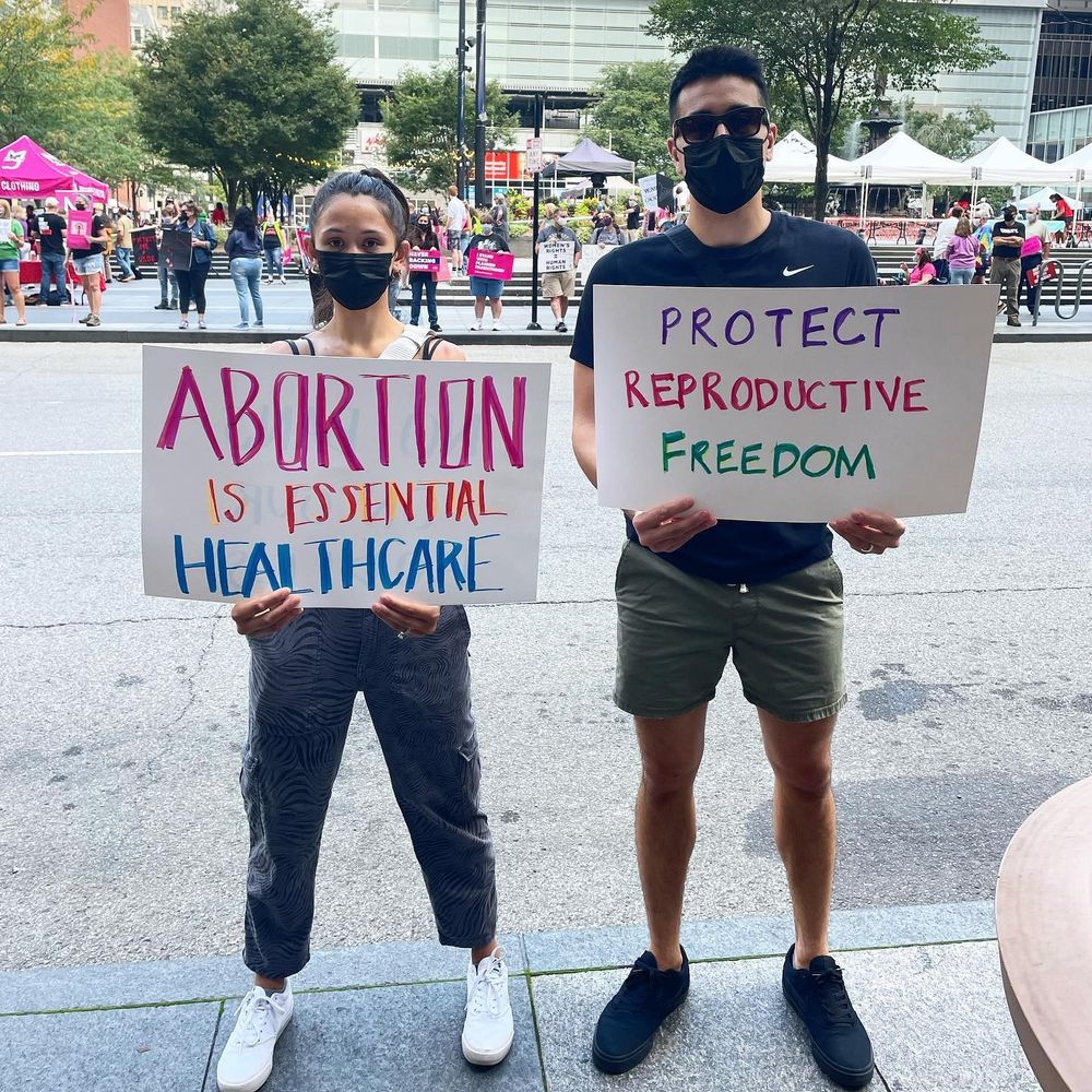 Two masked people at a protest. The woman is holding a sign reading ABORTION IS ESSENTIAL HEALTHCARE. The man is holding onee that reads PROTECT REPRODUCTIVE FREEDOM.