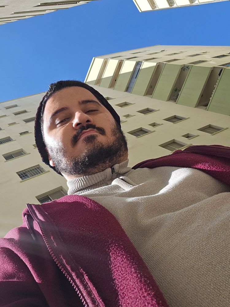Homem barbado com toca. Jaqueta de moletom vinho e camisa de lã. Céu azul atrás de prédios.