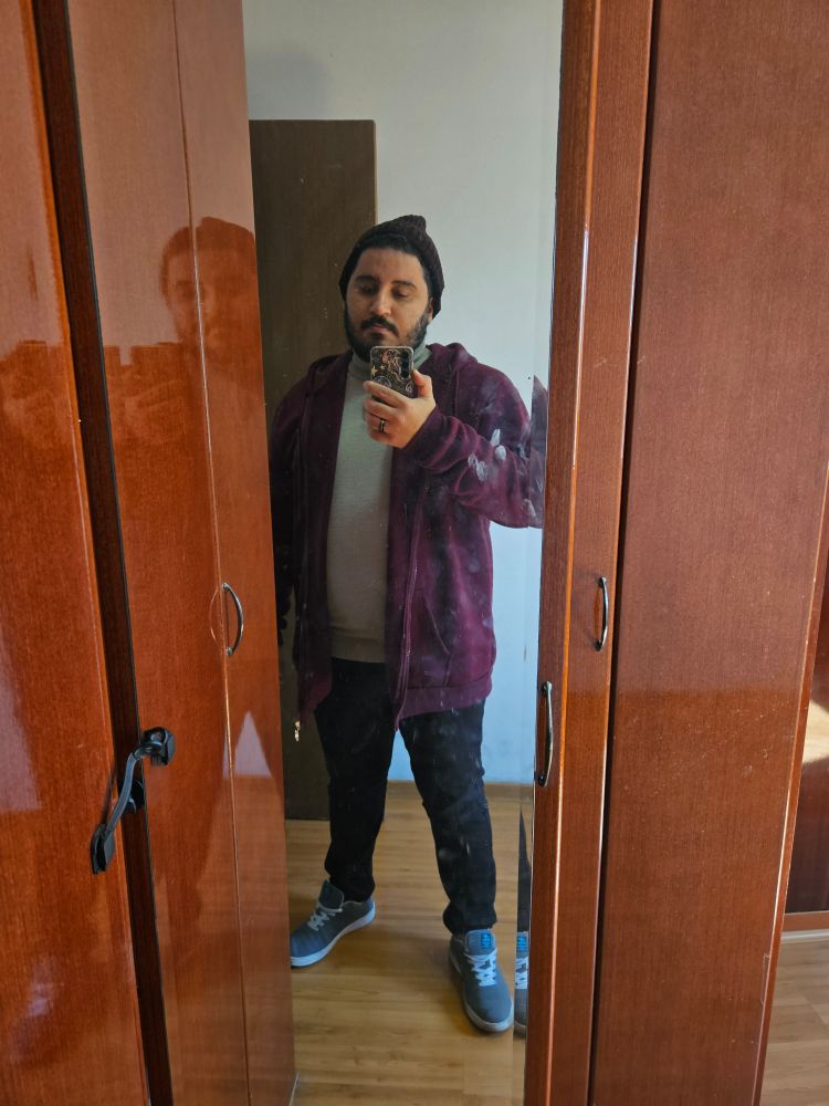 Homem barbado em frente a espelho de guarda roupa. Veste jaqueta de moletom vinho com uma camisa de lã por baixo, toca, calça preta e tênis cinza