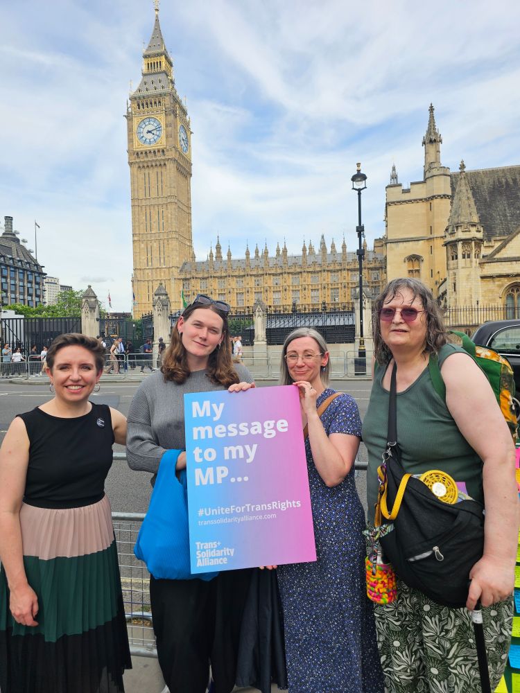 A group of 4 people stand in front of the Houses of Parliament. On the left is Carla Denyer MP. They are holding a placard in the colours of the trans flag which says "My message to my MP... #UniteforTransRights" 
