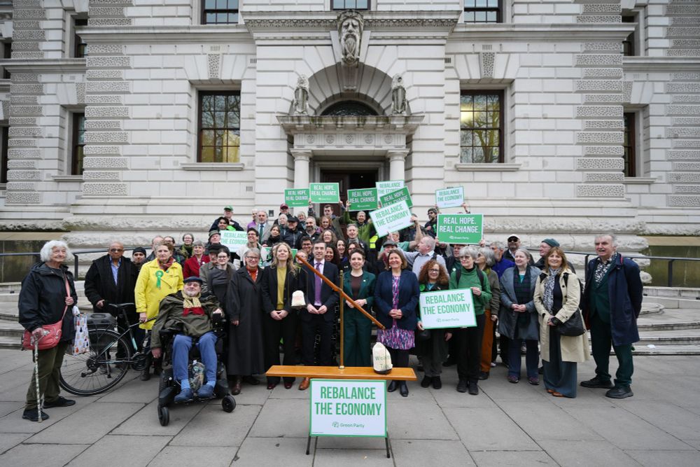 Green MPs Sian Berry, Adrian Ramsay, Carla Denyer and Ellie Chowns stand behind a giant set of scales. On one side is a heavy bag labelled 'wealth tax' and on the other side is a light bag labelled 'welfare cuts.' Behind the MPs is a crowd of Green Party supporters holding placards saying 'Real Hope, Real Change' and 'Rebalance the Economy'.
