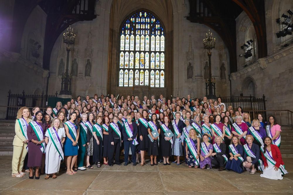 A group of women stand in an old building wearing green, white and purple sashes 
