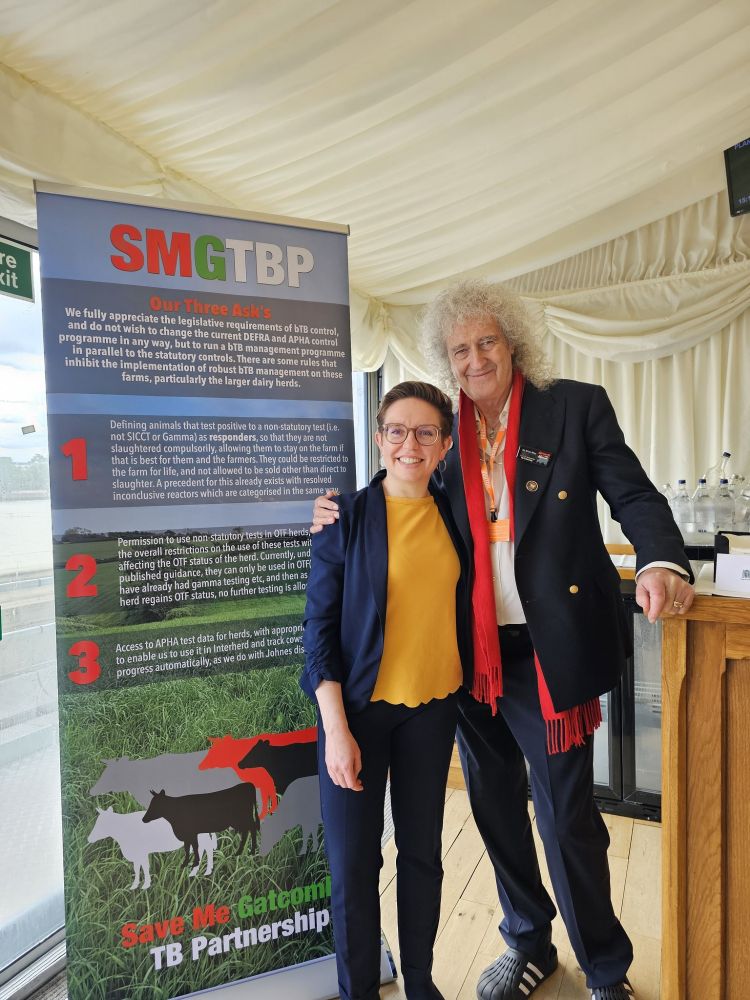 Carla and Brian May hug, standing in front of a banner with text outlining their three policy asks. 