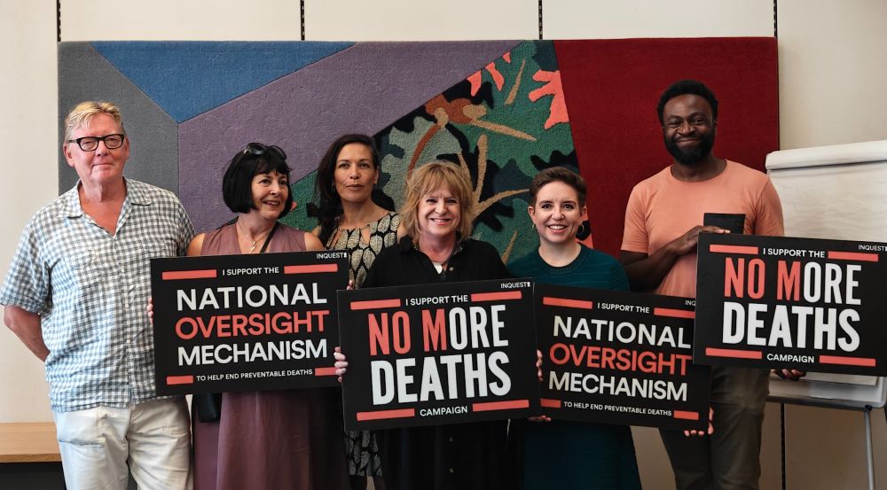 A group of people stand in front of a colourful wall, holding signs which say 'I support the National Oversight Mechanism' and 'No More Deaths.' Carla Denyer MP is in the group. 
