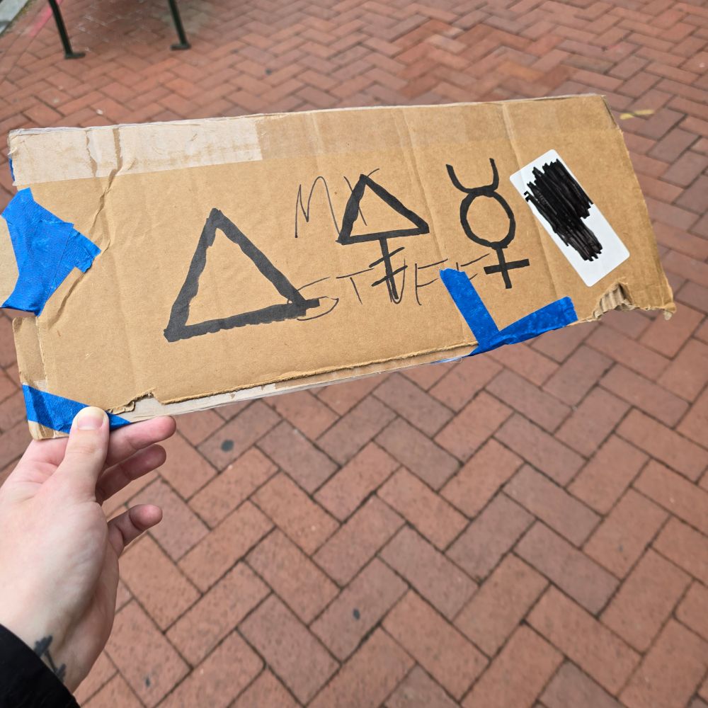The other side of the piece of cardboard. Smaller pieces of cardboard are held on with blue painters tape and a label from the box is blacked out with sharpy. In sharpie the alchemical symbols for Fire, Phosphorus, and Mercury are once again seen.