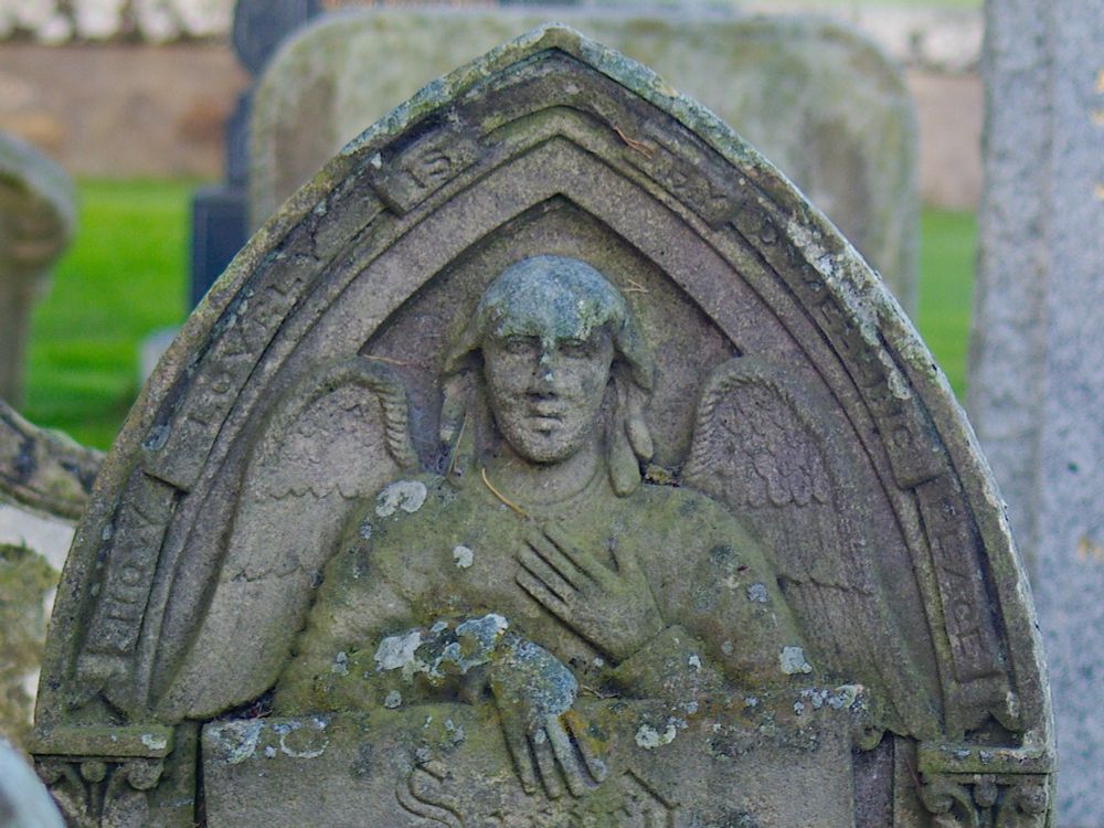 Carved head, shoulders and chest of an angel in low relief within the top of a pointed arch gravestone. In one hand the angel holds a scroll with the inscription. The other hand is held against the angel's breast. 