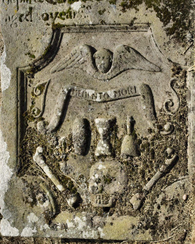 Close up of a panel from an outdoor grave slab of greenish-grey stone, with some moss covering parts and thatch from mowing. 

Within a rectangle with scalloped corners is carved - from top to bottom:
A cherub's head and wings.
A scroll, broadly resembling a draw-knife in shape, with the words Memento Mori, all between the initials I S in raised script lettering.
Below and partly between the ends of the scroll three emblems of mortality. Left to right a coffin, an hourglass, and a handbell.
Slightly lower and to the left and right of the first three emblems are, on the left an emblem that cannot be easily identified due to moss - possibly a trowel, and on the right a spade with handle at the top.
At the bottom is a large skull in profile between two long-bones in a V shape, the long bones and descending parts of the scroll forming a loose frame round the elements of mortality.


