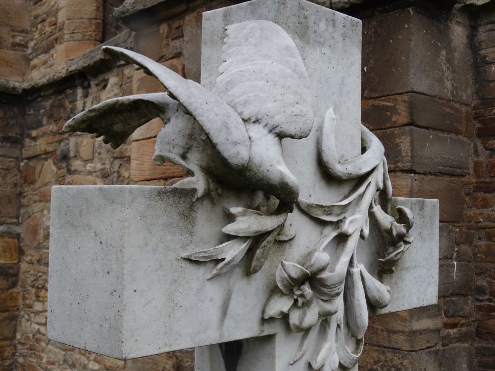 White marble cross with a carved dove perched on the left arm and pecking at lilies carved in high relief emerging from the upper arm and spreading across the face of the cross.