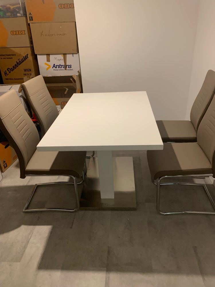 Auf dem Bild ist ein rechteckiger Esstisch mit weißer Tischplatte und stabilem Mittelfuß zu sehen. Rund um den Tisch stehen vier gepolsterte Stühle mit graubraunen Sitzflächen und Lehnen. Die Stuhlbeine sind aus Metall und haben eine freischwingende Form.