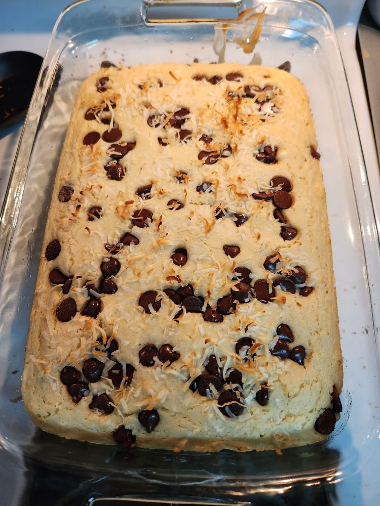 In a clear dish is a tan pancake that looks like a cake covered with brown chocolate chips and some white and brown coconut flakes 