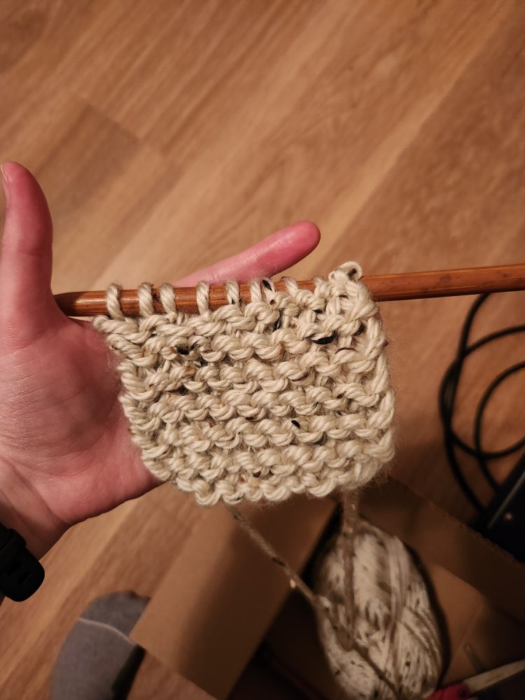 A white hand holding a wooden knitting needle with some off white knit on it. 