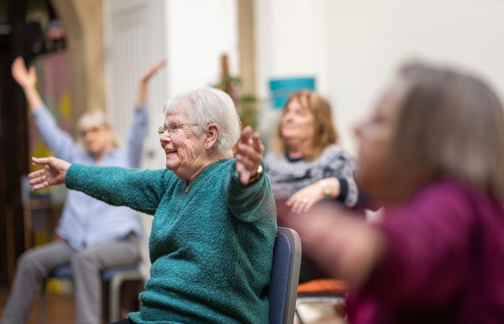 A group of older people in a exercise class on chairs waving their arms in the air and smiling. One person is in focus, the others are a little blurry. 