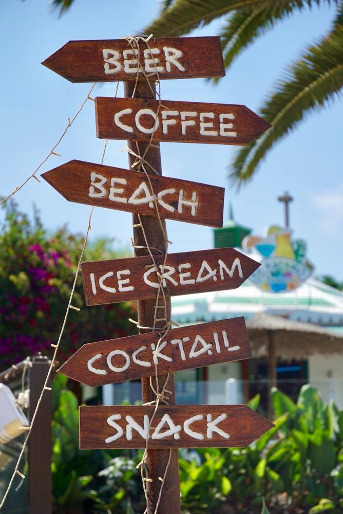 Braune Schilder untereinander an einem Pfahl mit Aufschriften. Im Hintergrund ein verschwommenes Gebäude und Pflanzen