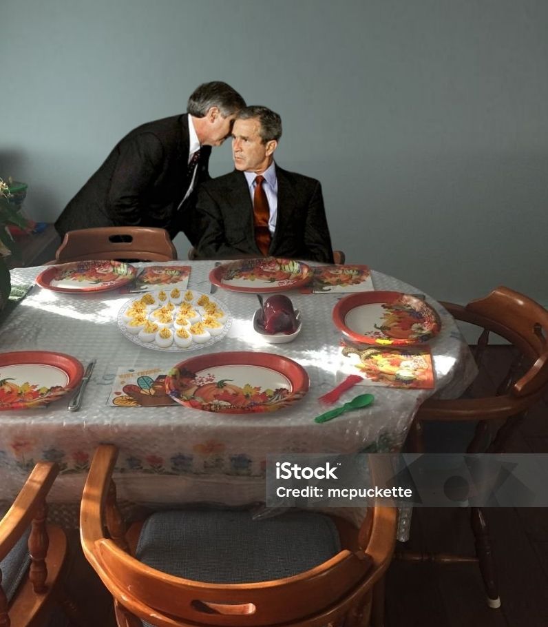 A man is leaning in and whispering to George W. Bush as he's sitting at an empty thanksgiving table. There's a plate of deviled eggs in the middle of the table. 