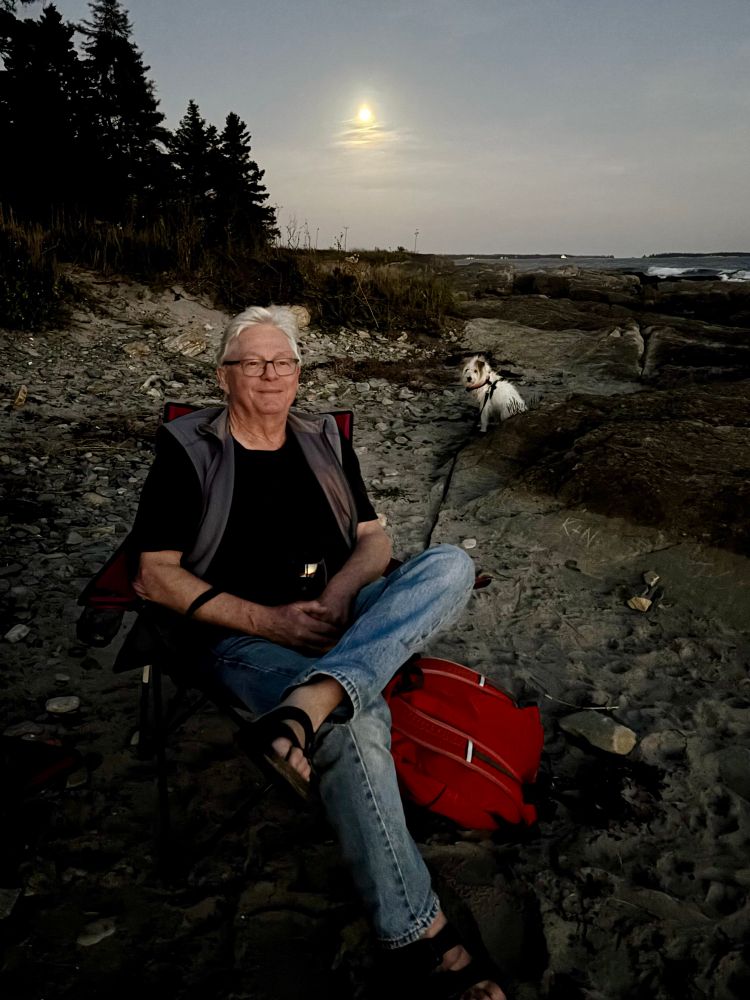 A grey haired man and small white dog are sitting on a small beach beside the ocean at dusk. A full moon rises in the sky behind them.