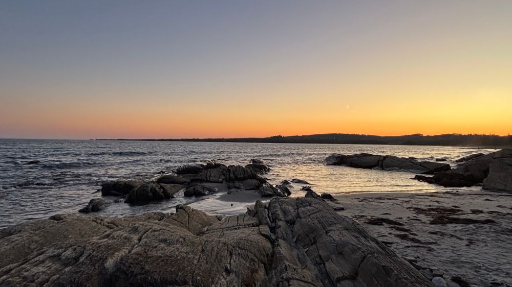 An orange, pink and yellow sky visible above the ocean. In the foreground, there’s a rocky shoreline with the same colours reflected in the water. 